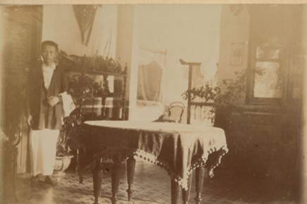 This photo shows a Vietnamese young Indigenous person in what looks to be a nice house of a French person. The boy looks to be a servant and is standing in the dining room of the French quarters.